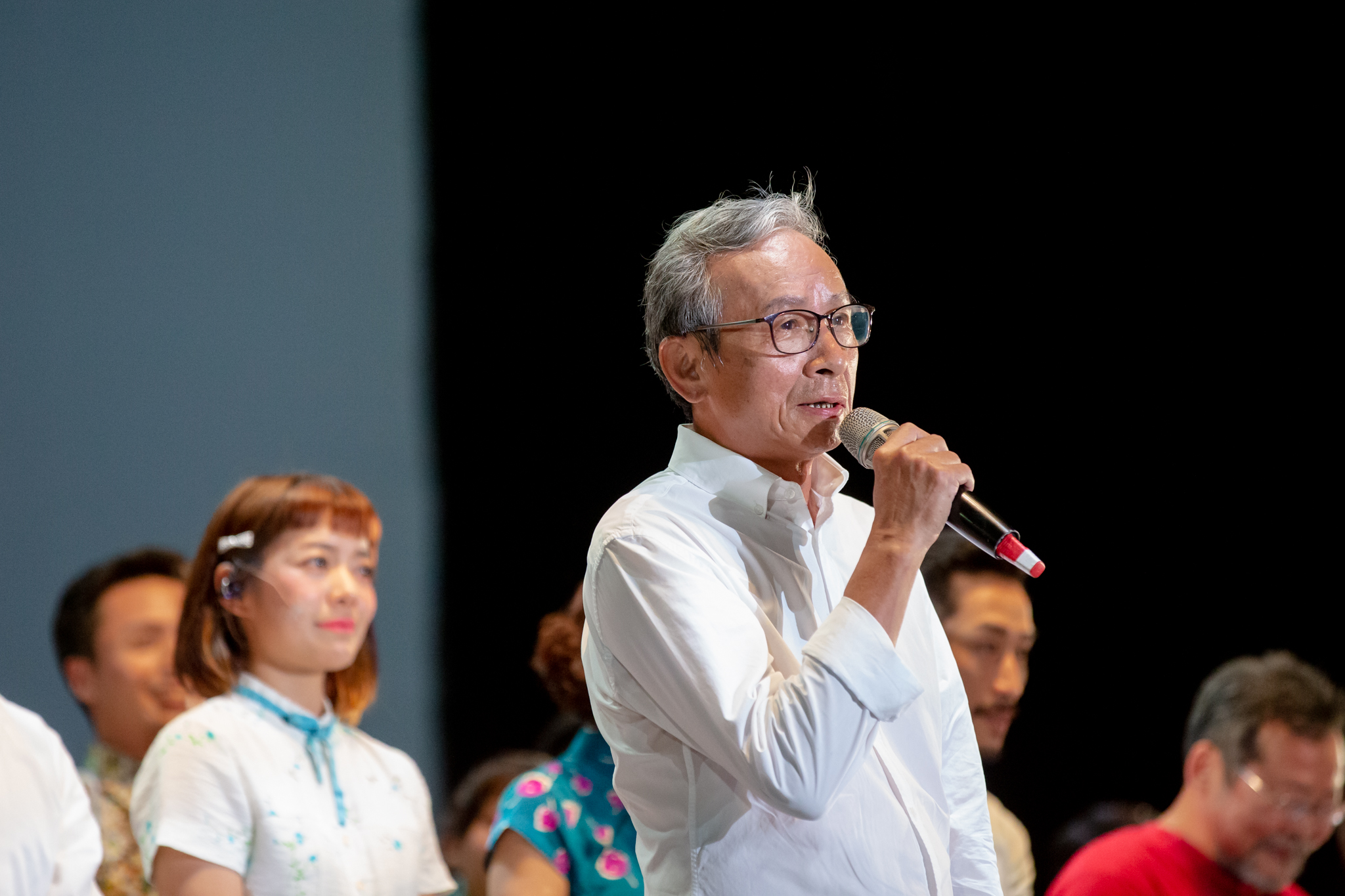 吳念真穿白襯衫、戴眼鏡,手拿麥克風在台上致詞